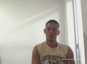 En la foto está un hombre moreno, cabellos cortos crespos, orejas y nariz grandes, vestido con una camisilla beige y se encuentra sentado al interior de una oficina asistiendo virtualmente a la audiencia en la que se le procesó. 