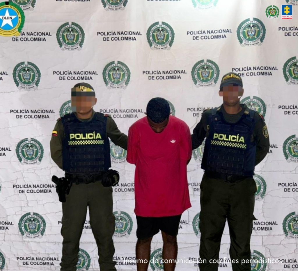 En la imagen está un hombre moreno, de pie, de frente con la cabeza agachada, brazos atrás esposados, de baja estatura, vestido con camiseta roja y bermuda negra. Se encuentra acompañado por dos policías.