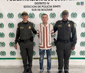  En la foto está un hombre blanco, de pie, de mediana edad, de frente, con los brazos atrás esposados, tiene el cabello negro corto, está vestido con una camisa beige y rayas café, jean azul y tenis. A su lado se encuentran dos policías. 