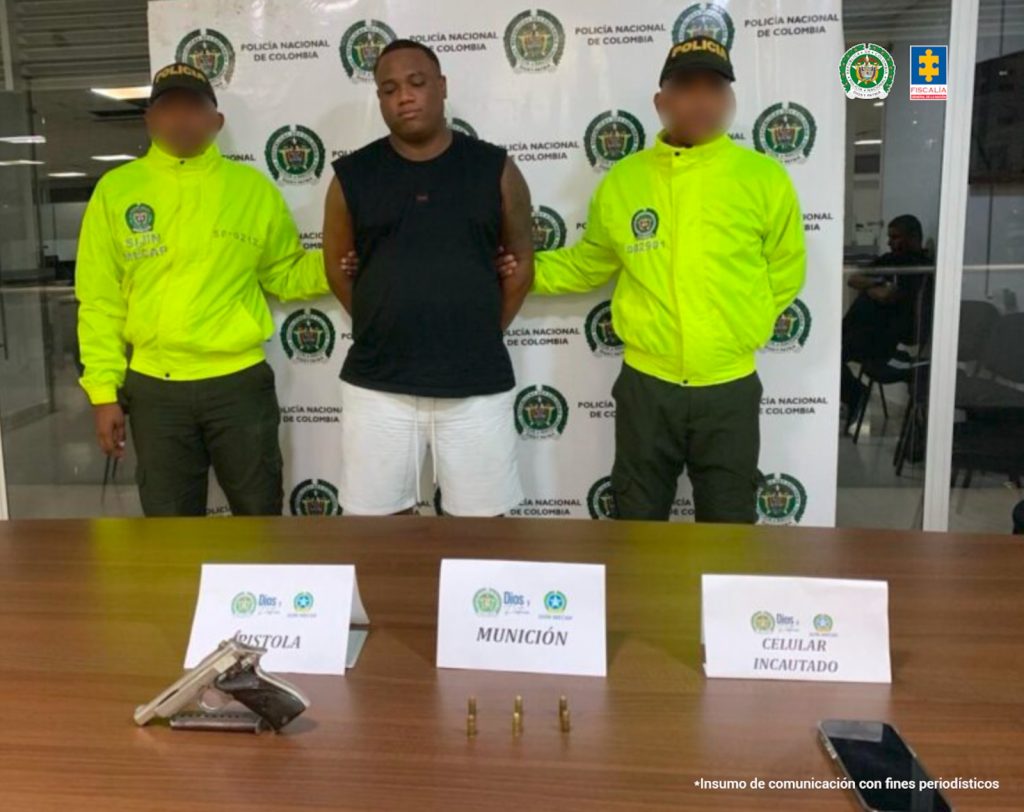 En el cuadro hay un hombre de pie, con los brazos atrás, de contextura gruesa, moreno, vestido de camisilla negra y bermuda blanca, custodiado por dos agentes de la Policía Nacional. Delante de ellos está una mesa donde están el arma y las municiones incautadas.