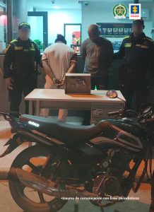 En la foto se encuentran dos hombres, uno vestido de camiseta y pantalón blanco, y el otro de camiseta gris con jean, custodiados por dos servidores de la Policía Nacional. En una mesa está el arma incautada y al lado la motocicleta decomisada.