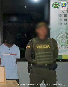 En la fotografía esta un hombre delgado con la cabeza agachada y los brazos atrás, vestido de camiseta blanca y pantalón negro acompañado por un policía.