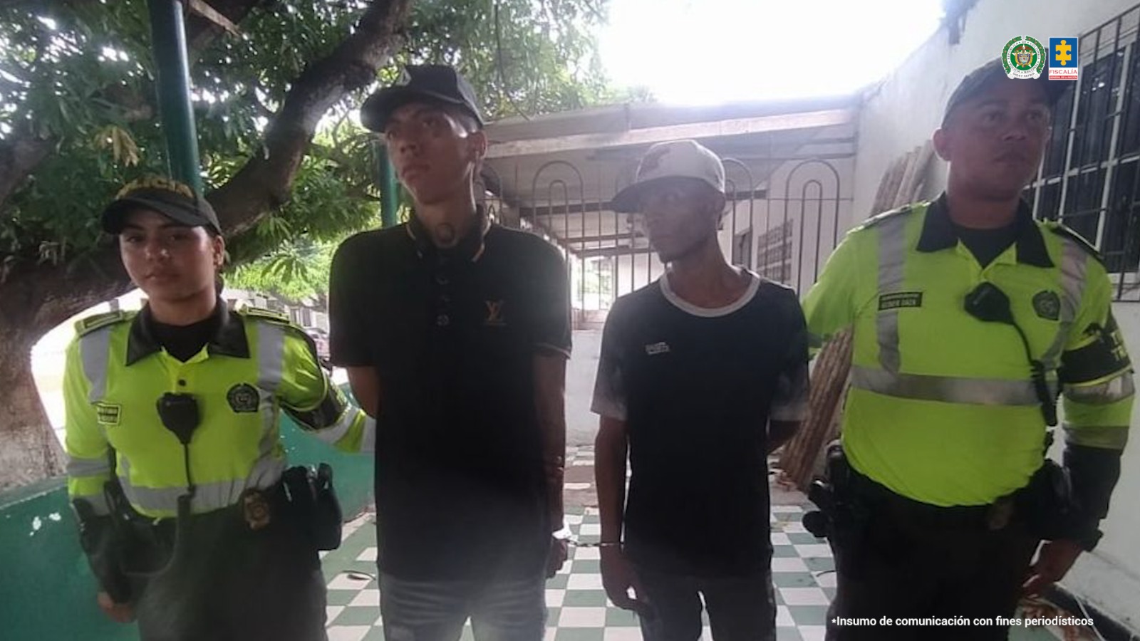 En la fotografía están dos hombres morenos de pie, de frente, delgados, con los brazos esposados, ambos tienen camisetas negras, uno tiene gorra negra y el otro gorra blanca y se encuentran custodiados por dos uniformados de la Policía Nacional. 