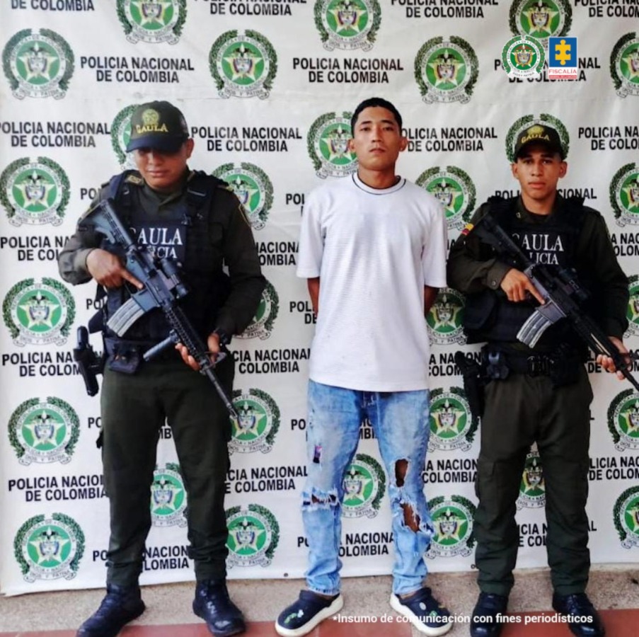 En la foto se encuentra un hombre trigueño de pie con los brazos atrás esposados, cabellos cortos negros, vestido con una camista gris, jean azul y zapatos oscuros.A su lado se encuetran dos uniformados de la Policía Nacional.