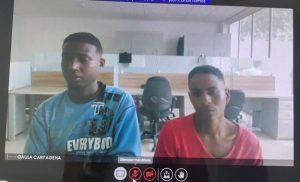 En la foto están los medios cuerpos de dos hombres morenos, uno vestido con camiseta azul y el otro con camiseta roja. Se encuentran sentados al interior de una oficina asistiendo virtualmente a la audiencia en la que se le procesó. 