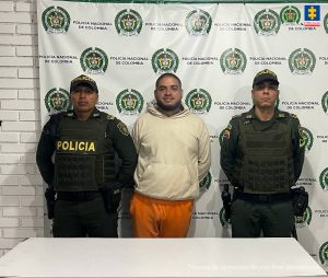 En la fotografía se aprecia de pie y con los brazos atrás a Carlos Andrés Rodríguez Ángel. Viste buso beage y pantalón color naranja. A sus extremos hay uniformados de la Policía Nacional. Delante de ellos hay una superficie blanca y detrás un pendón de la Policía.