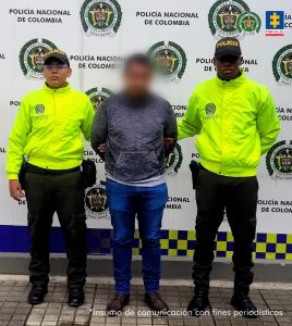 En la fotografía se aprecia de pie, con los brazos atrás al judicializado. Viste buso de manga larga gris, jean zul y zapatos negros. A sus extremos hay uniformados de la Policía Nacional. Y detrás de ellos hay un pendón institucional de la Policía.