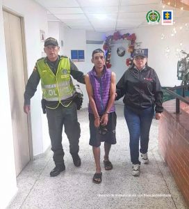 En la fotografía se aprecia en acción de caminata por un pasillo a Héctor Fabio Gómez Mosquera. Lleva los brazos adelante, esposado y tiene una gorra oscura en las manos. Viste camisa de manga ziza lila, bermuda negra y pantuflas oscuras. Al lado derecho de él hay un uniformado de la Policía Nacional y al lado izquierdo una servidora del CTI.