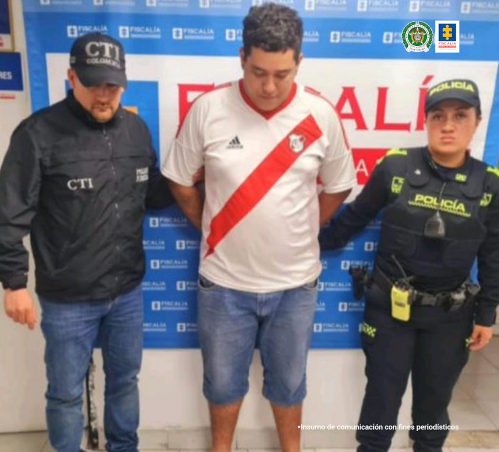 En la fotografía se aprecia de pie y con los brazos atrás a Jharvinn Santiago Quintero Ruiz. Viste camisa blanca de manga corta con franja roja diagonal en el frente, cuello y hombros; asimismo, bermuda de jean azul. Al lado derecho de él hay un servidor del CTI de la Fiscalía y al lado izquierdo una uniformada de la Policía Nacional. Detrás de ellos hay un pendón institucional de la Fiscalía.