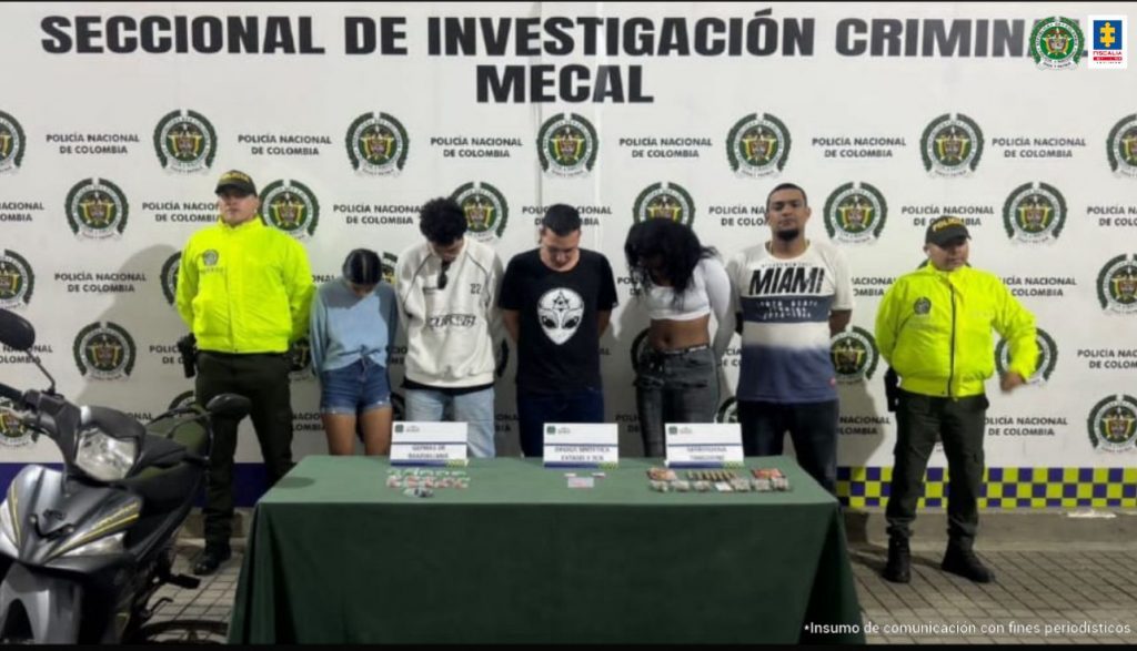 En la fotografía se aprecia a los presuntos integrantes del grupo delincuencial Los Bananeros, de pie y con los brazos atrás. A lado y lado de ellos hay uniformados de la Policía Nacional. Delante de ellos hay una mesa con mantel verde y sobre ella se aprecia varias papeletas de plástico y tres letreros. Detrás de ellos hay un pendón institucional de la Policía.