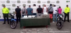 En la fotografía se aprecia de espaldas a la cámara a los presuntos integrantes del grupo delincuencial Los Del Hoyo. A lado y lado de ellos hay uniformados de la Policía Nacional. También se aprecia una mesa con mantel verde y sobre ella varios elementos y dos motocicletas. En la parte posterior hay un pendón institucional de la Policía.