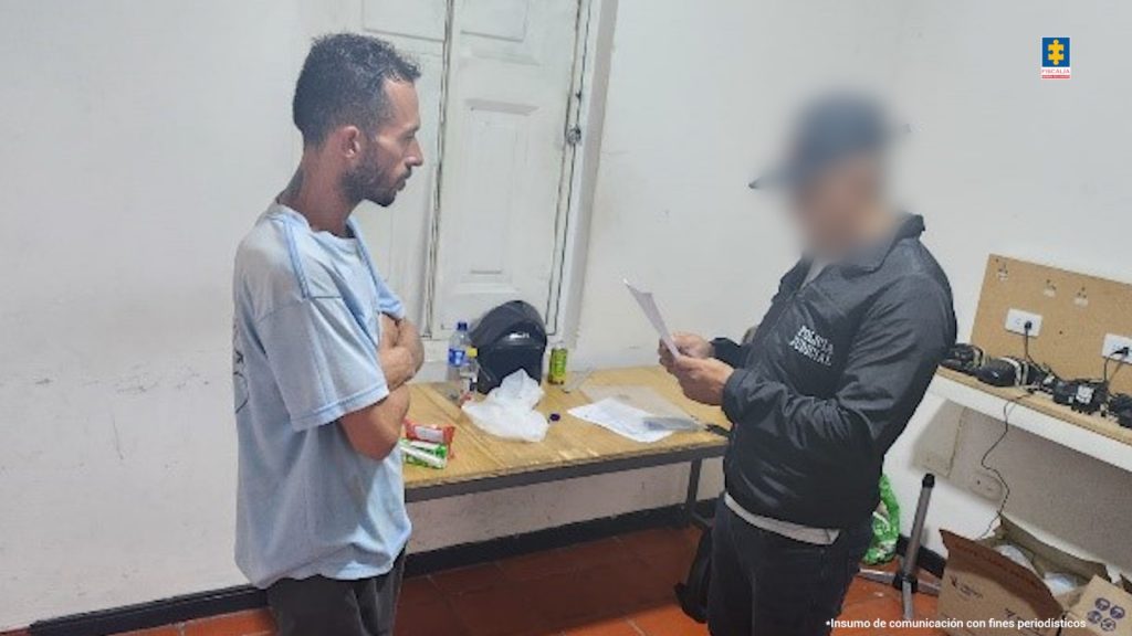 En la fotografía se aprecia de pie, con los brazos entrecruzados, de lado a la cámara, dentro de un sitio cerrado a Róbinson Laverde. Viste camiseta de manga corta de color azul claro; bermuda oscura y pantuflas. Frente a él hay un servidor del CTI de la Fiscalía en acción de lectura de un documento.