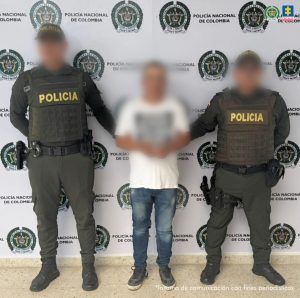 El capturado se encuentra de frente y custodiado por dos uniformados de la Policía Nacional. Detrás de la foto hay un backing de la Policía y el capturado tiene un buzo blanco, jean y tennis negros.