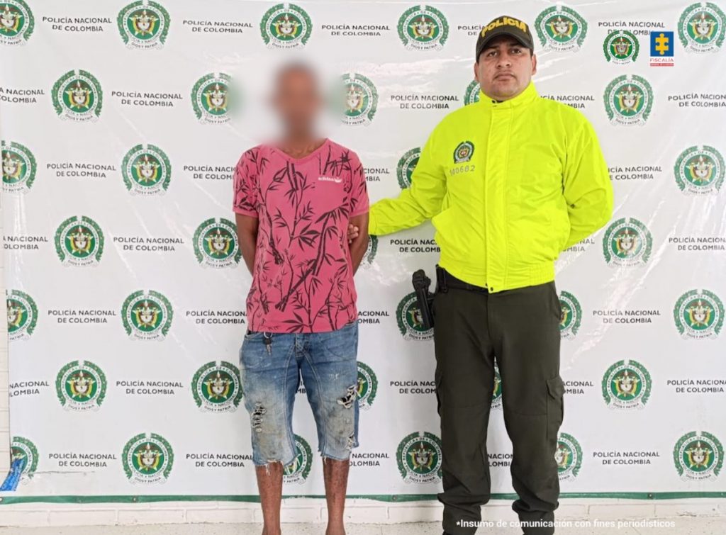 En la imagen aparece una persona de suéter rojo y pantaloneta de jean, junto con un uniformado de la Policía.