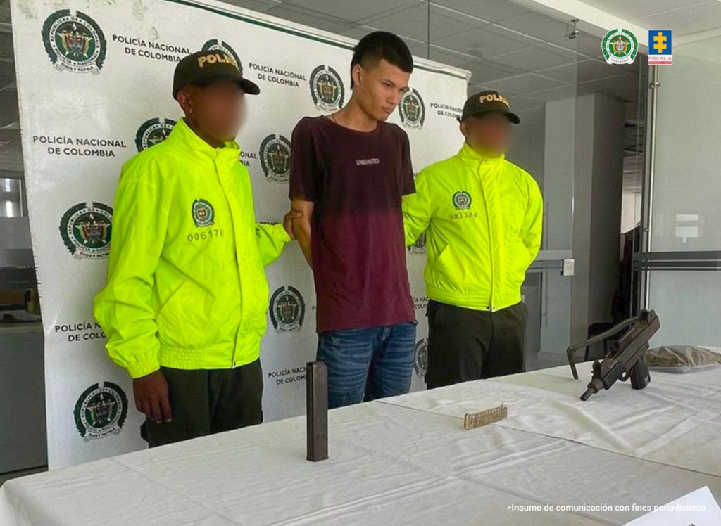 En la imagen está un hombre de pie, con los brazos atrás, delgado, vestido con camiseta vino tinto y jean azul, custodiado por dos agentes de la Policía Nacional. Delante de ellos hay una mensa con el arma y la droga incautada.