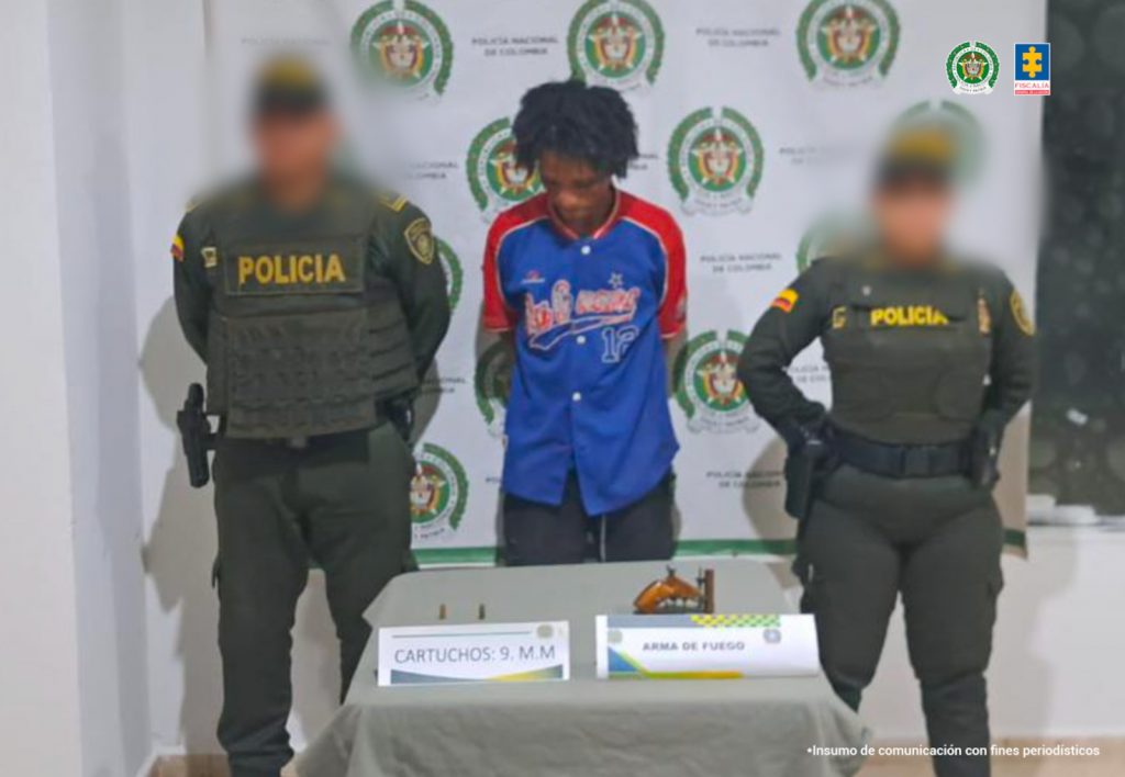En la fotografía, aparece un hombre de pie, moreno, delgado con la cabeza agachada de cabellos largos crespos, vestido de camiseta azul con rojo y pantalón negro. Se encuentra acompañado de dos uniformados de la Policía Nacional.