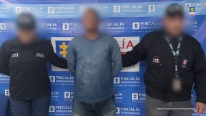 En la imagen está un hombre moreno, de pie, con los brazos atrás, de contextura gruesa, vestido de buzo y jean azul, con el rostro difuso, custodiado por dos funcionarios del CTI. Detrás de ellos hay un pendón azul de la Fiscalía.