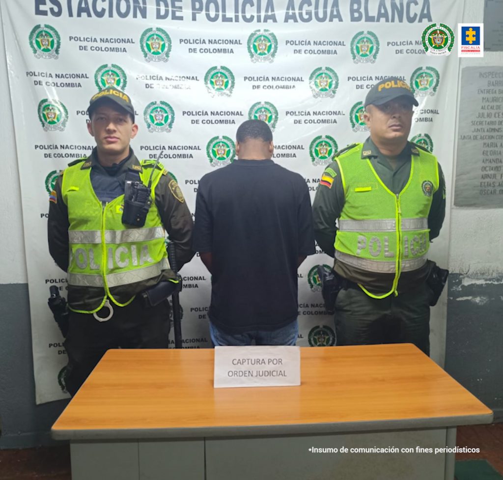 En la fotografía se aprecia a Jhon Jader Valencia Castillo, de pie y de espaldas a la cámara. Viste camiseta de manga corta de color negro y jean azul. A lado y lado de él hay uniformados de la Policía Nacional. En la parte delantera hay una superficie de color madera y base gris, y sobre ella hay un letrero que dice: ‘Captura por orden judicial’. En la parte posterior hay un pendón institucional de la Policía.