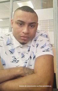 En la fotografía está el rostro de un hombre joven, trigueño, de ojos y cabellos negros, nariz y labios gruesos, vestido de camisa blanca con azul. Se encuentra sentado en la audiencia donde se le procesó.