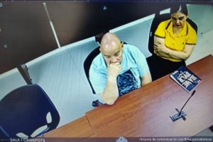  En la fotografía aparece un hombre sentado con los brazos sobre un escritorio de tez blanca, calvo y vestido de camiseta azul, con un micrófono en su boca.  A su lado se encuentra una mujer sentada de cabello negro, vestida con camiseta amarilla y pantalón negro