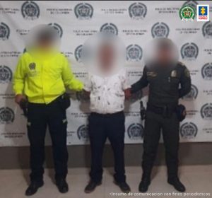 En la fotografía se encuentra un hombre con el rostro difuminado, de pie, de estatura baja vestido con camisa blanca, pantalón y zapatos negros, custodiado por dos uniformados de la Policía Nacional. Detrás de ellos hay un pendón de esa institución.