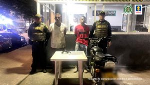 En la imagen aparecen dos hombres morenos, uno vestido con camiseta blanca y pantalón beige y el otro con camiseta roja y jean. Ellos se encuentran custodiados por dos uniformados de la Policía Nacional. También hay una mesa blanca donde está el arma que se les incautó.