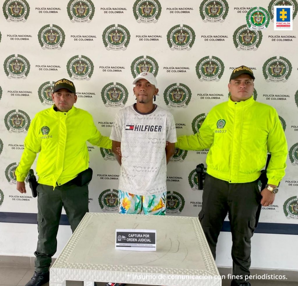 En la fotografía hay un hombre delgado, moreno, con los brazos atrás, vestido de camiseta blanca, gorra blanca y bermuda estampada, custodiado por dos agentes de la Policía Nacional. Detrás de ellos hay un pendón de la Policía Nacional.