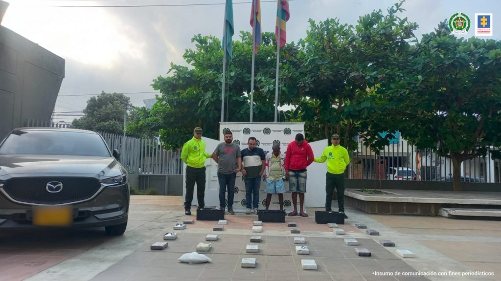 En la fotografía aparecen cuatro hombres de pie, custodiado por dos agentes de la Policía Nacional. Delante de ellos está la droga incautada y detrás de ellos un pendón de esa institución.