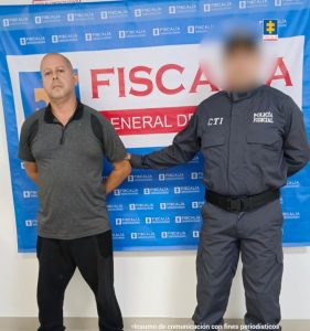 En la fotografía aparece un hombre de pie, con los brazos atrás, de tez blanca, calvo, vestido con una camiseta gris y pantalón negro. Detrás de ellos hay un pendón de la Fiscalía.