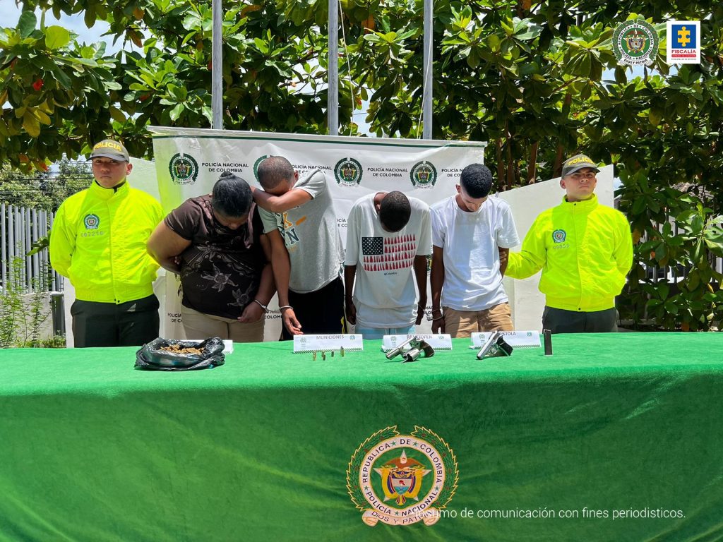 En la fotografía se observan a tres hombres y una mujer con las cabezas agachadas, custodiados por dos agentes de la Policía Nacional. Delante de ellos hay una mesa con mantel verde donde están las armas y las drogas incautadas.