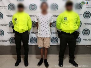 En la fotografía hay un hombre moreno de pie, con los brazos atrás, vestido con camiseta blanca, bermuda beis, zapatos tenis negros, custodiado por dos agentes de la Policía Nacional. Detrás de ellos hay un pendón de esa institución.