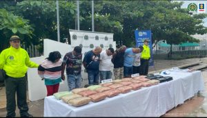 En la fotografía están seis hombres y una mujer de pie, con la cabeza agachada, custodiados por dos uniformados de la Policía Nacional. Delante de ellos hay una mesa blanca que tiene encima la droga incautada.