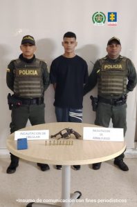 En la fotografía está un hombre joven, de pie, de tez trigueña, con los brazos atrás, vestido con una camiseta negra y un jean azul. Está custodiado por dos uniformados de la Policía Nacional.