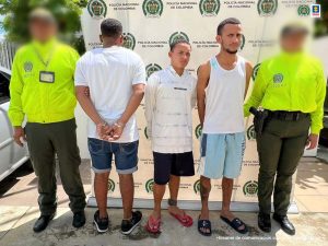 En la fotografía aparecen tres hombres vestidos de camisetas blancas con bermudas, todos se encuentran de pie, custodiados por dos agentes de la Policía Nacional. Detrás de ellos hay un pendón de esa institución.
