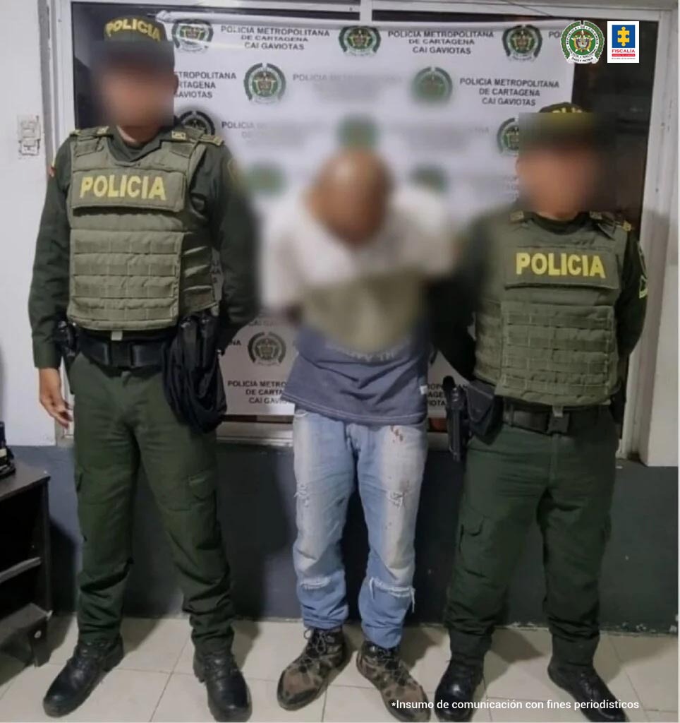 En la fotografía se observa a un hombre con la cabeza agachada y con los brazos atrás, vestido de camiseta azul, jean azul y botas negras, custodiado por dos agentes de la Policía Nacional. Detrás de ellos aparece un pendón de esa institución.