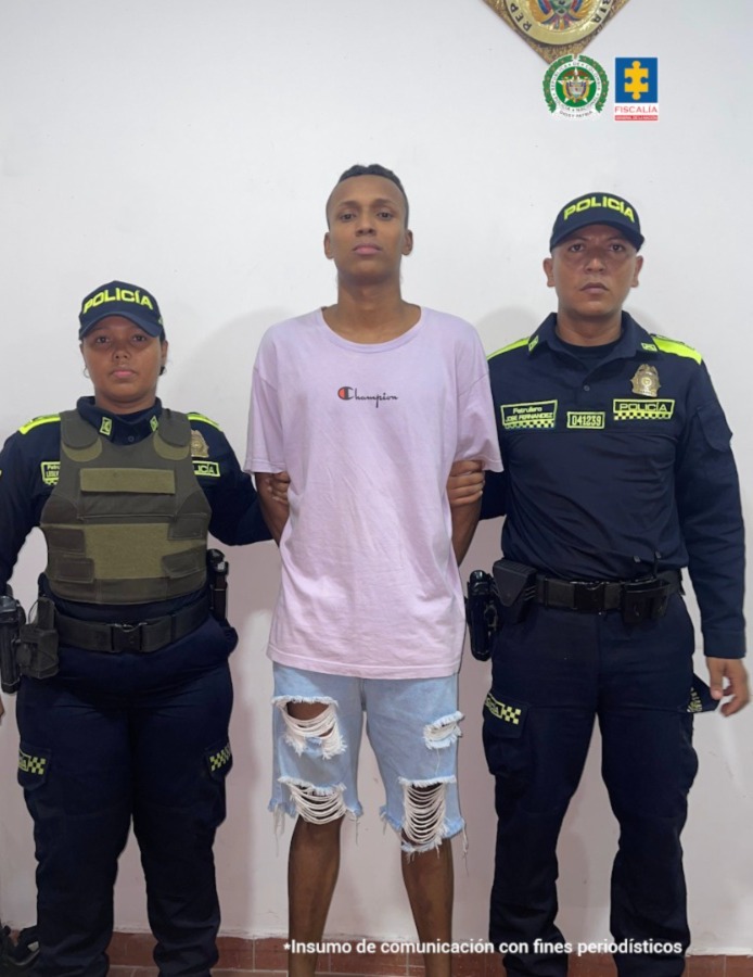 En la fotografía está un hombre de pie, alto, moreno, delgado, cabellos cortos negros, vestido con camiseta morada, bermuda de jean azul y sandalias negras. Se encuentra custodiado por dos uniformados de la Policía Nacional.