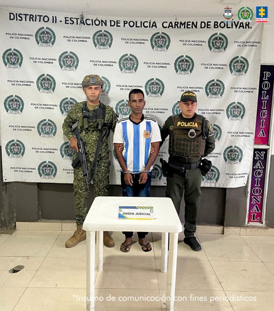 En la foto está un hombre de pie, moreno, estatura baja con los brazos adelante esposados, vestido con una camiseta blanca y rayas azules, un jean azul oscuro y unas sandalias negras. Se encuentra custodiado por un uniformado de la Policía Nacional y otro de Infantería de Marina.