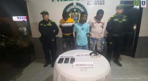 En la imagen se observa a tres hombres de tez morena custodiados por dos agentes de la Policía Nacional. Delante de ellos se encuentra una mesa con el arma incautada, documento falso y celulares.