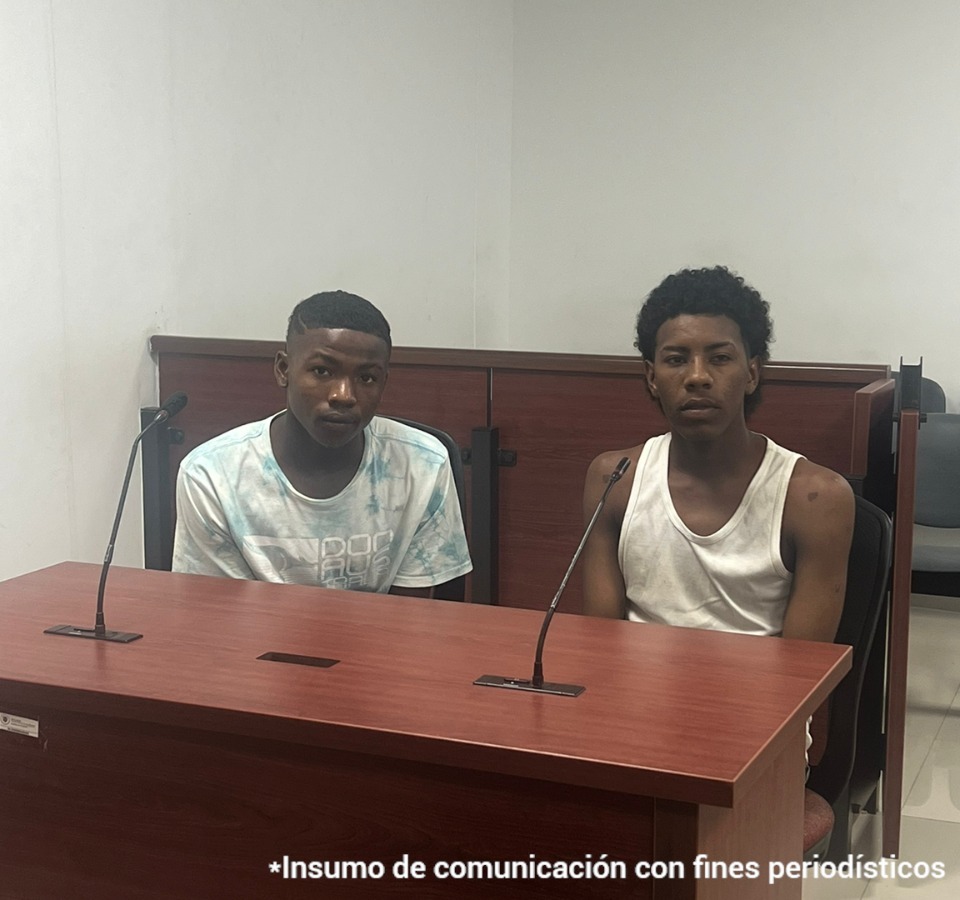 En la imagen aparecen dos hombres morenos, ambos con camisetas blancas, sentados con micrófonos frente a su boca durante la audiencia ante el juez de control de garantías.