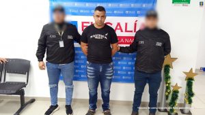 En la fotografía está un hombre joven de pie, de frente, trigueño, cabellos negros cortos, con los brazos atrás esposados, vestido con un jean azul, camiseta y zapatos negros y se encuentra custodiado por dos uniformados del CTI. Detrás de ellos hay un pendón de la Fiscalía. 