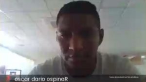 En la fotografía está el rostro de un hombre de tez morena, cabellos negros se encuentra sentado en una oficina, escuchando virtualmente la audiencia donde es procesado y aparece vestido con una camiseta blanca