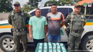 En la fotografía aparecen dos hombres de pie, uno de estatura baja vestido de camiseta verde con jean negro y el otro de alta estatura vestido con camiseta gris y jean azul. Se encuentran custodiados por dos agentes de la Policía Nacional. Delante de ellos hay una mesa con el dinero incautado.
