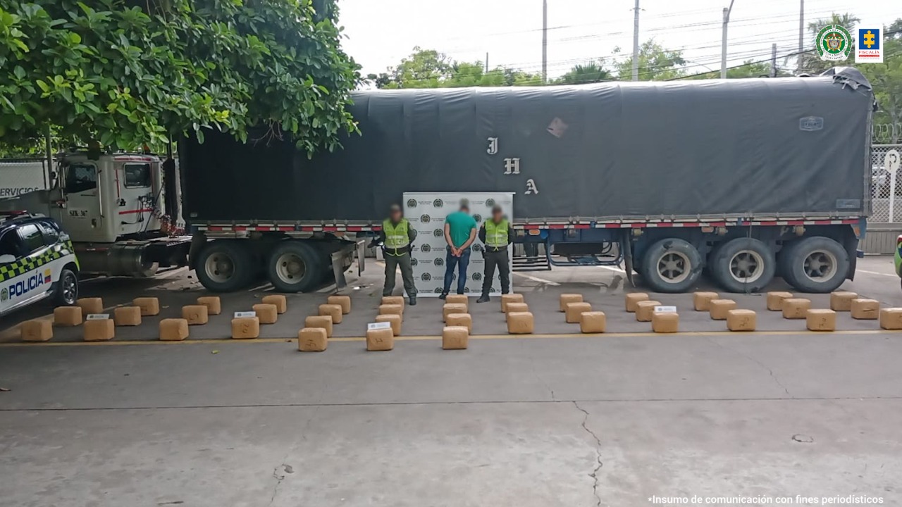 En la fotografía se observa a un hombre de camiseta verde de espalda custodiado por dos agentes de la Policía Nacional y varios paquetes en el suelo con el camión donde presuntamente se transportaba la droga.