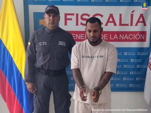 En la fotografía aparece un hombre moreno, de baja estatura, con los brazos adelante esposados, medianamente grueso, vestido con camiseta blanca y pantalón blanco, custodiado por un uniformado del Cuerpo Técnico de Investigación. Detrás de ellos hay un pendón azul de la Fiscalía.