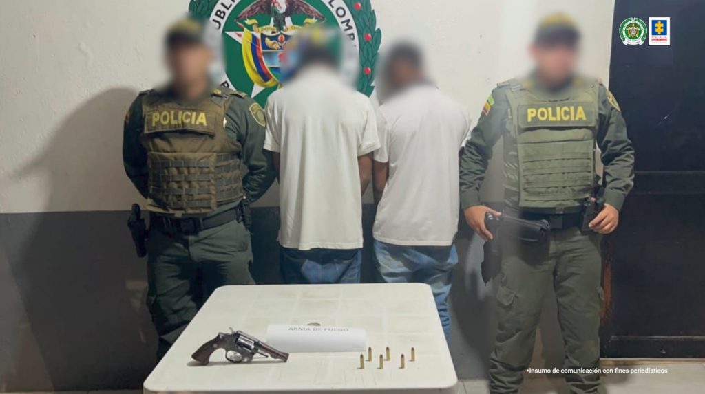 En la fotografía aparecen dos hombres de espalda vestidos con camiseta blanca y jean, custodiados por dos uniformados de la Policía Nacional. Detrás de ellos hay un pendón de esa institución y delante de ellos se encuentra una mesa con mantel verde que tiene en la parte superior un arma de fuego.
