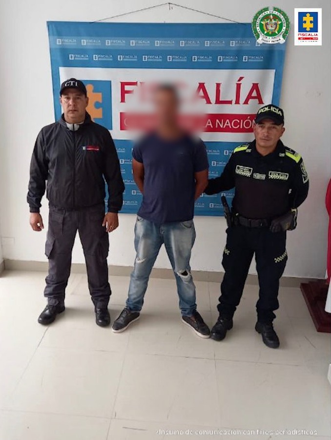 En la imagen aparece a un hombre de camiseta azul oscura con jean, custodiado por un uniformado del CTI de la Fiscalía y por un funcionario de la Policía Nacional. Detrás de ellos se encuentra un pendón de la Fiscalía.