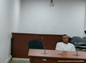  En la foto está el medio cuerpo de un hombre moreno, joven, de cabellos cortos crespos, se encuentra sentado en una silla frente a un escritorio de madera con micrófono, vestido con una camiseta blanca asistiendo a la audiencia donde se le procesó.