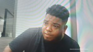 En la fotografía aparece el medio cuerpo de un hombre moreno, joven, de contextura gruesa, labios y nariz grande, pelo crespo, vestido con una camiseta negra, se encuentra sentado escuchando la audiencia en la que se le procesó.