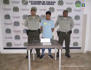 En la fotografía esta un hombre de pie con los brazos atrás, vestido de camiseta azul clara, jean azul, tenis negro, custodiado por dos uniformados de la Policía Nacional. Delante de ellos hay una mesa con la cadena recuperada.