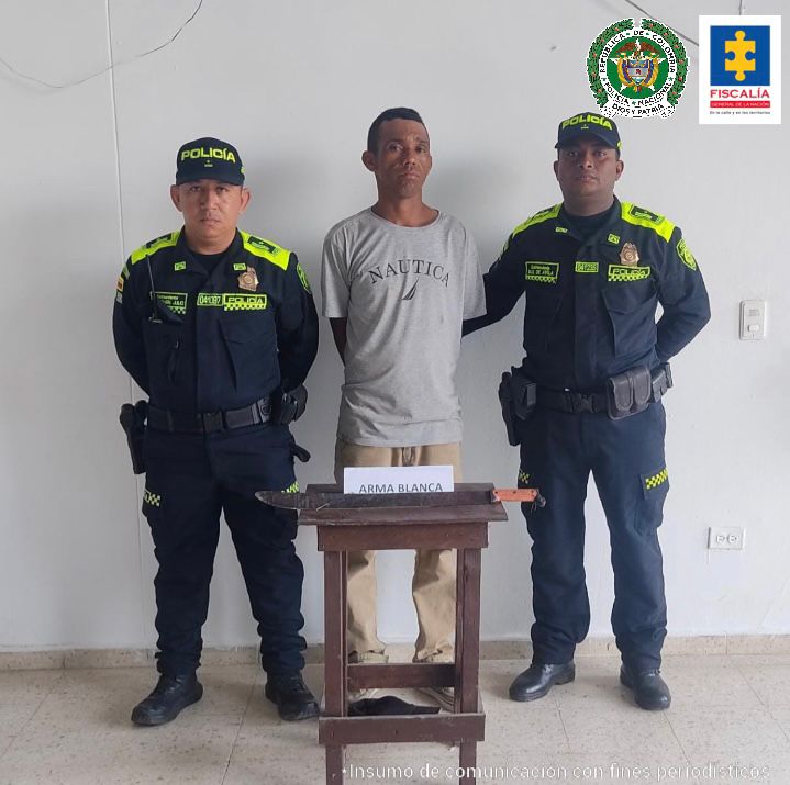 En la imagen se observa a un hombre de camiseta gris con pantalón beis, custodiado por dos uniformados de la Policía Nacional. Delante del capturado se divisa una mesa que tiene en la parte superior el arma blanca incautada.